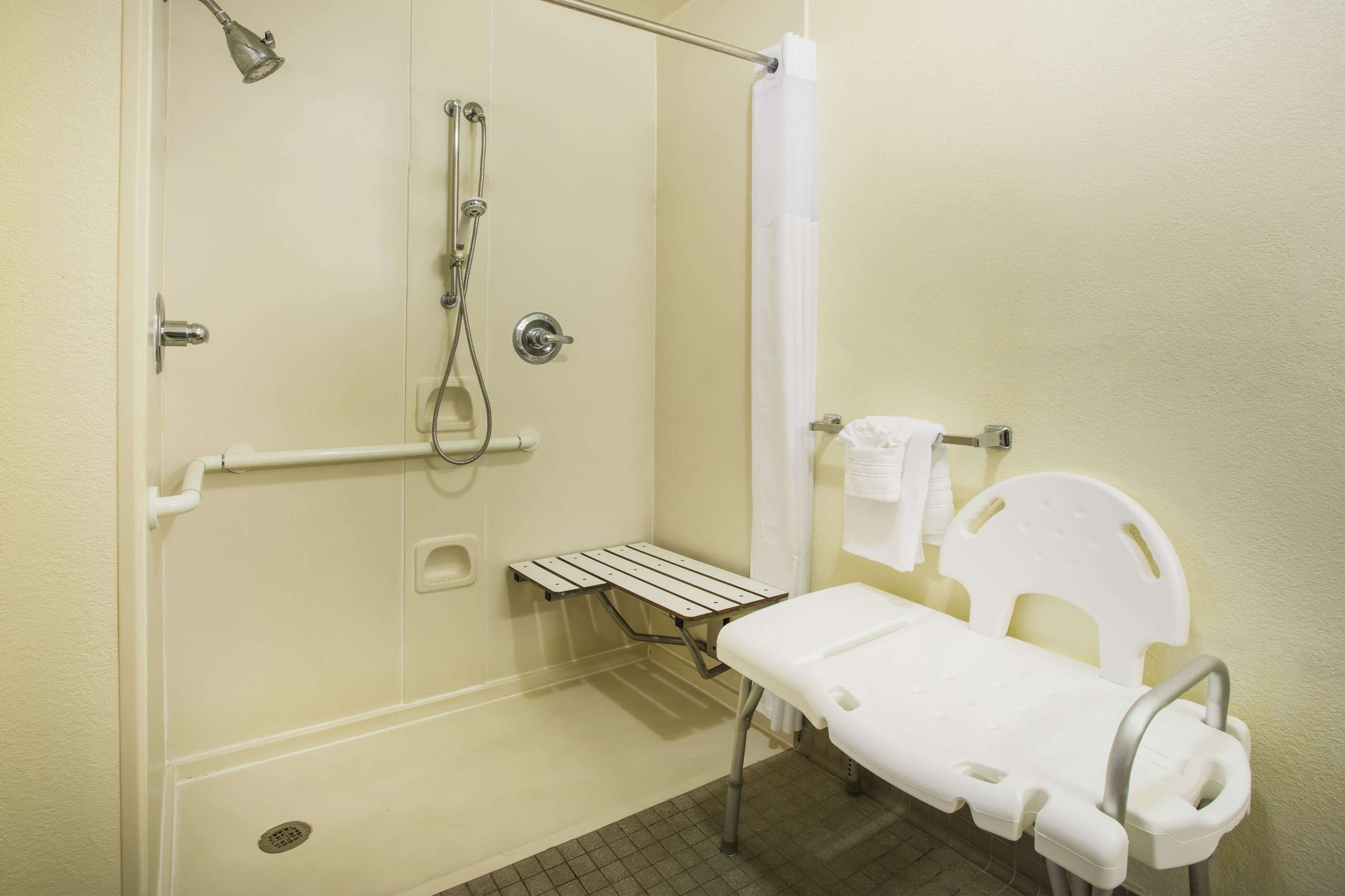 A bathroom with a shower, a grab bar, a white toilet, and a bench.