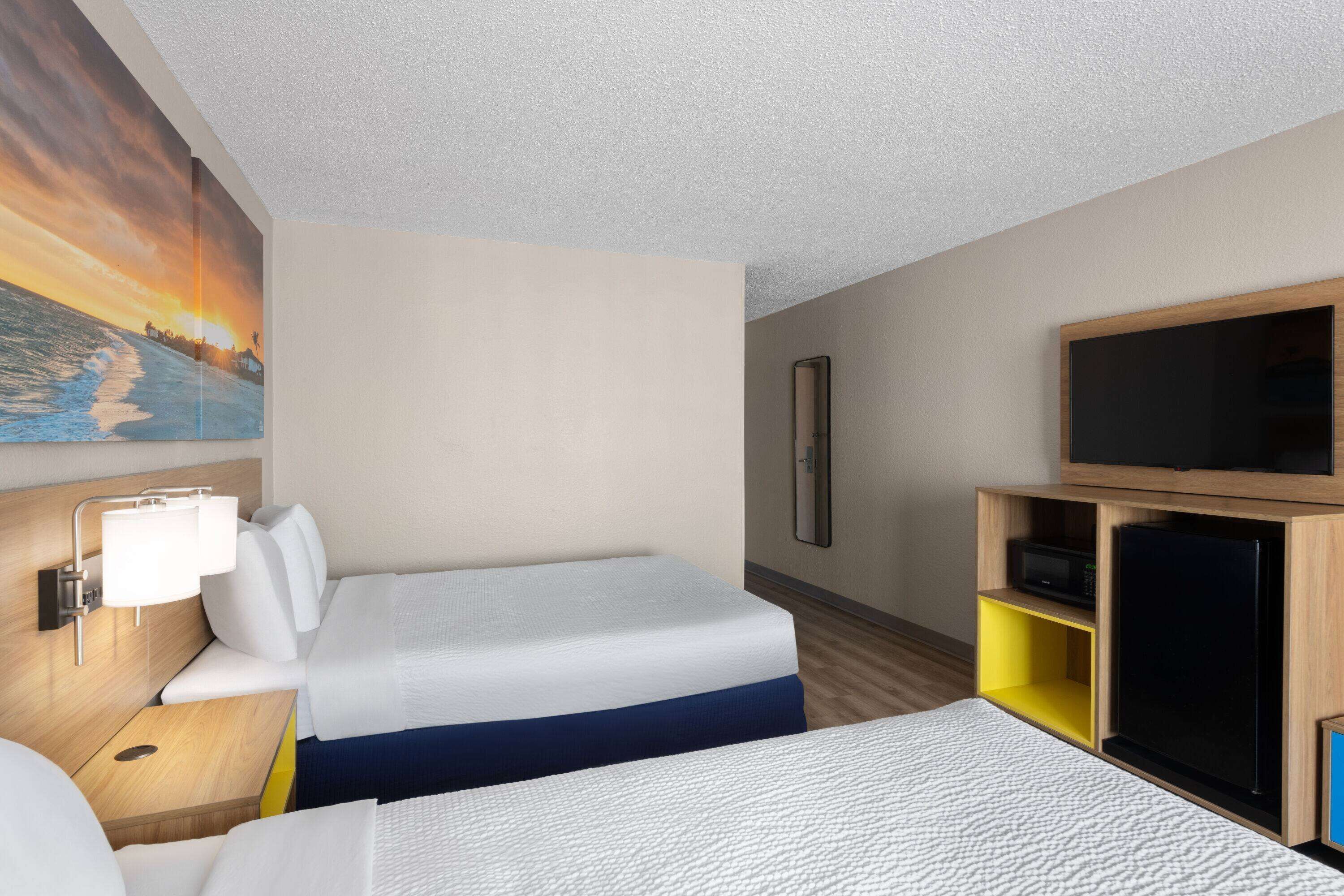 A hotel room with two beds, a TV, a desk with a lamp, and a painting of a sunset.