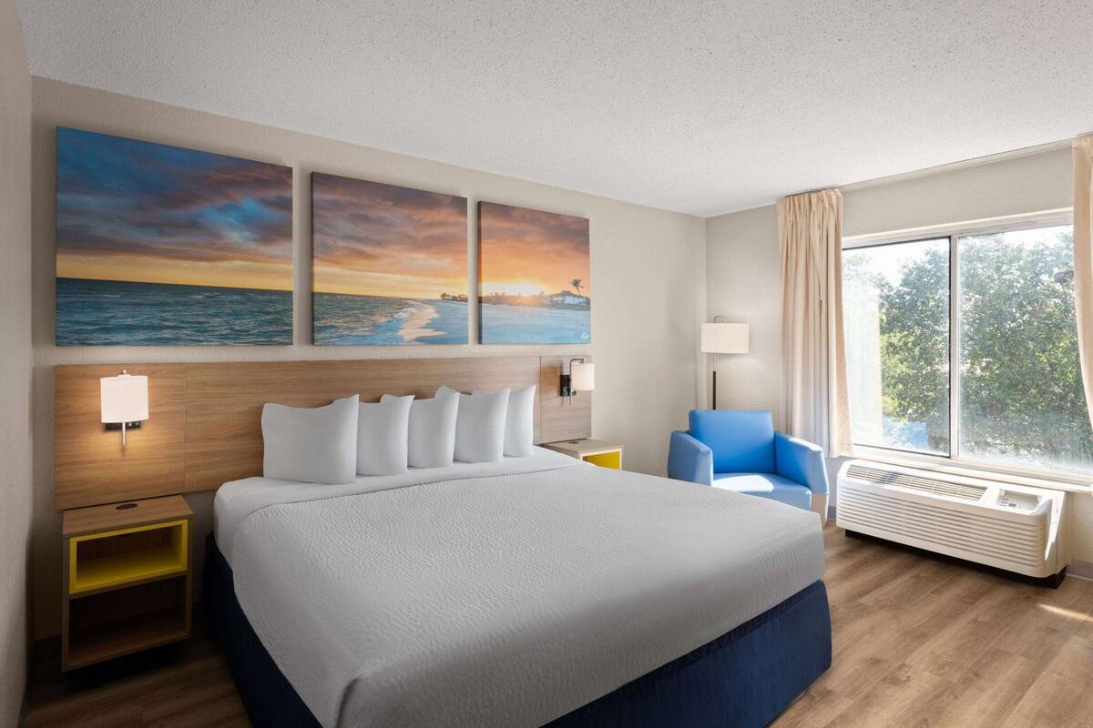 A hotel room with a large bed, a nightstand, a desk, a chair, a window with curtains, and three paintings on the wall.