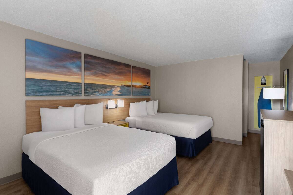A hotel room with two beds, a wooden headboard, a nightstand with lamps, and a wall-mounted artwork depicting a sunset over the ocean.