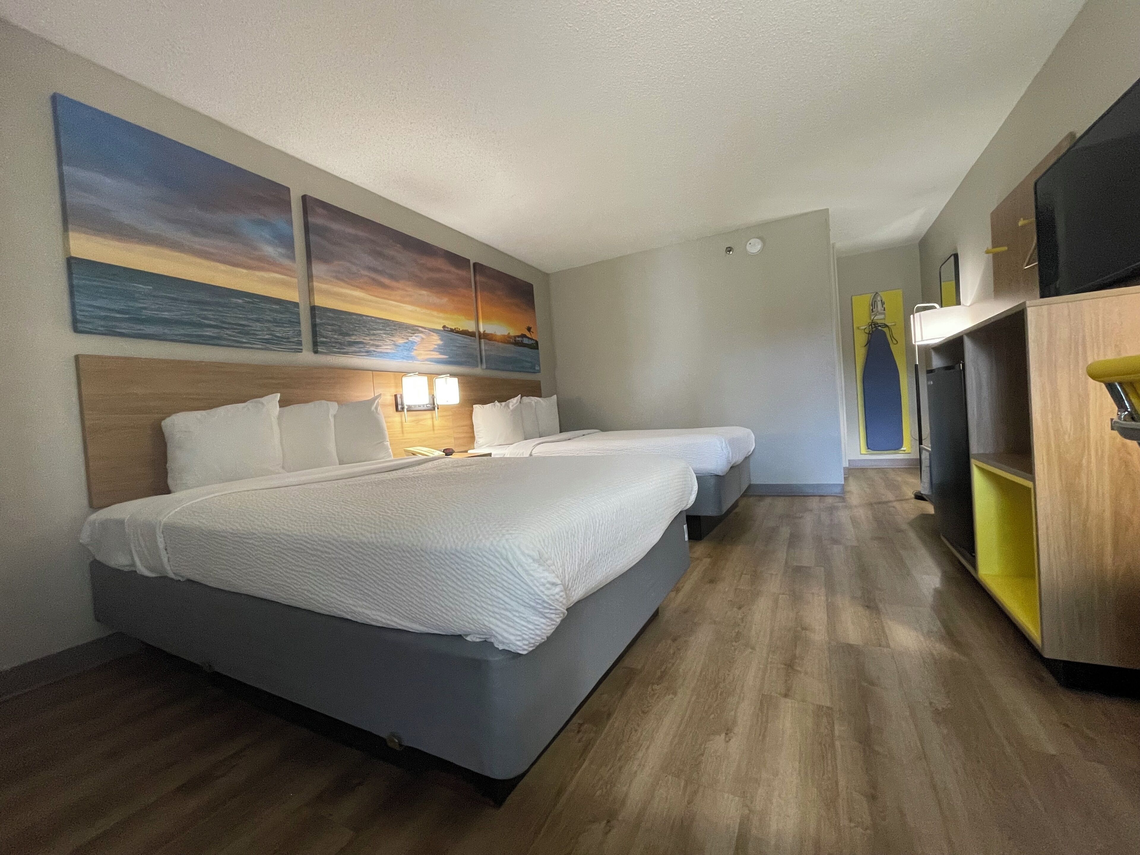 A hotel room with two beds, a wooden headboard, a wall-mounted TV, and a small kitchenette.
