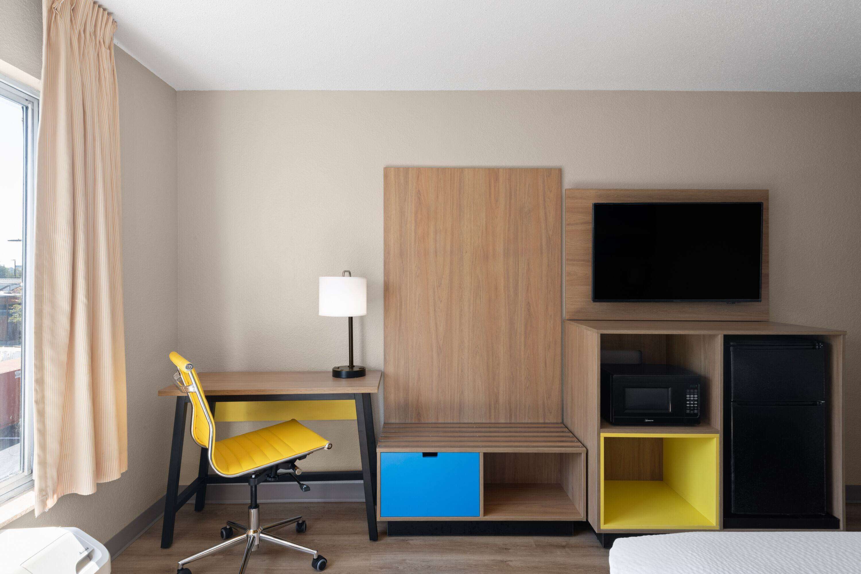 Une chambre d’hôtel avec un bureau en bois, une chaise jaune, un téléviseur, un four à micro-ondes, un réfrigérateur et un tiroir de rangement bleu.