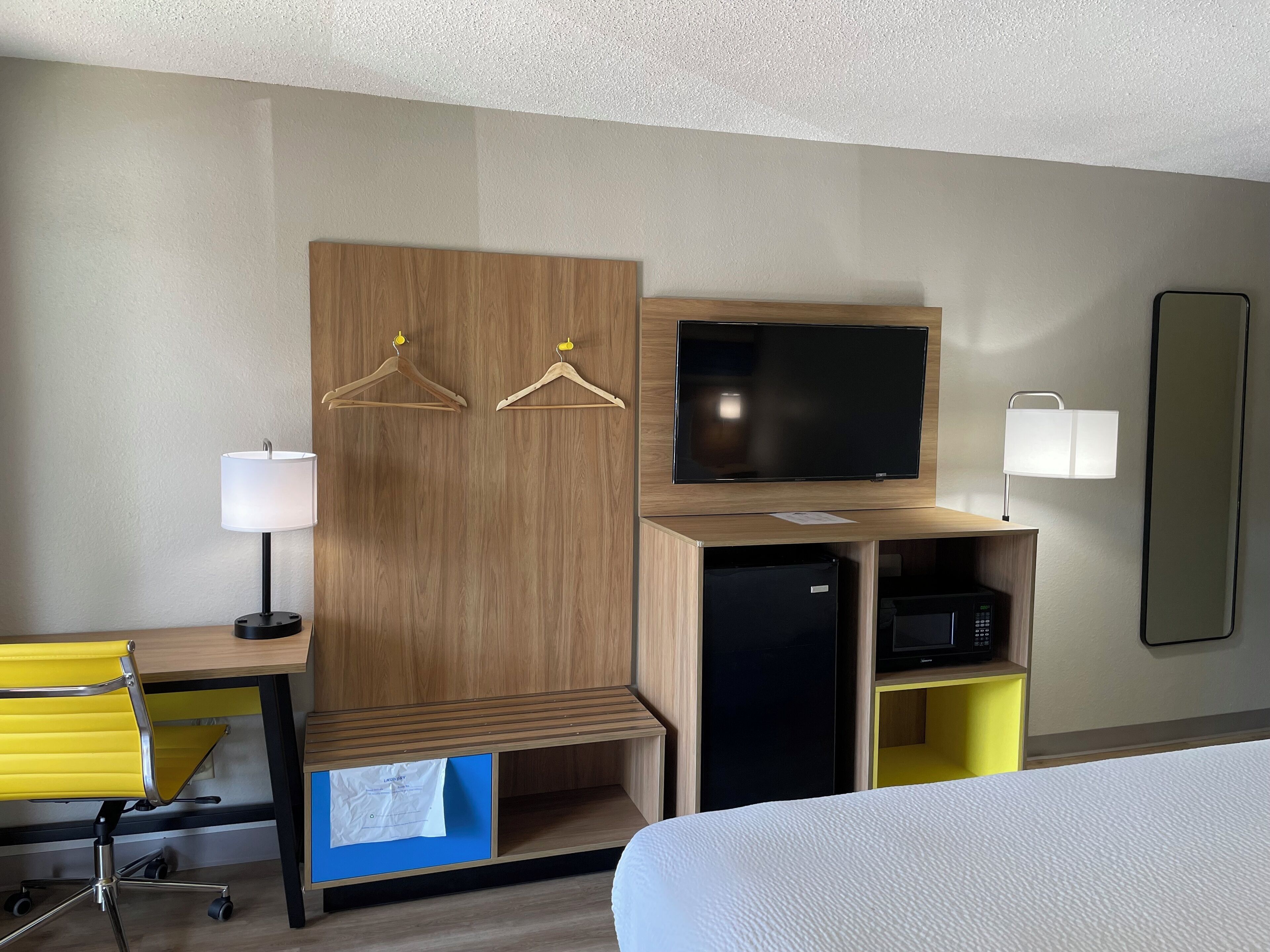 A hotel room with a wooden wardrobe, a flat-screen TV, a desk with a lamp, a yellow chair, a bed, and a microwave.