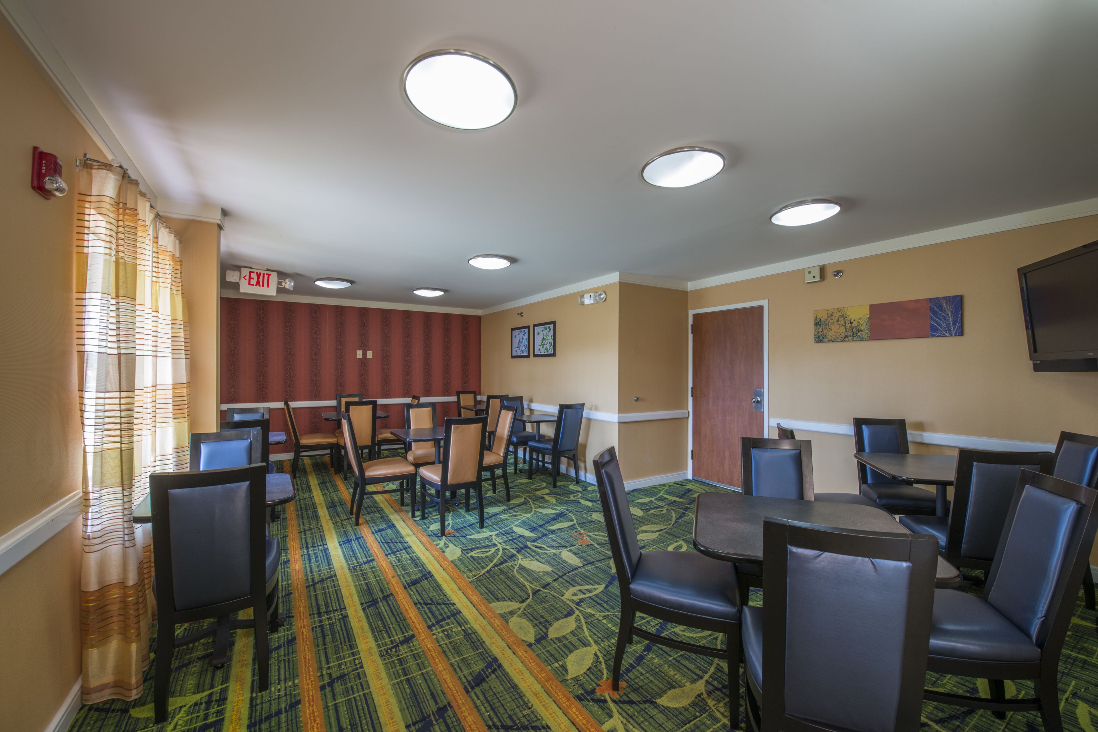 A conference room with a patterned carpet, multiple tables and chairs, ceiling lights, and an exit sign.