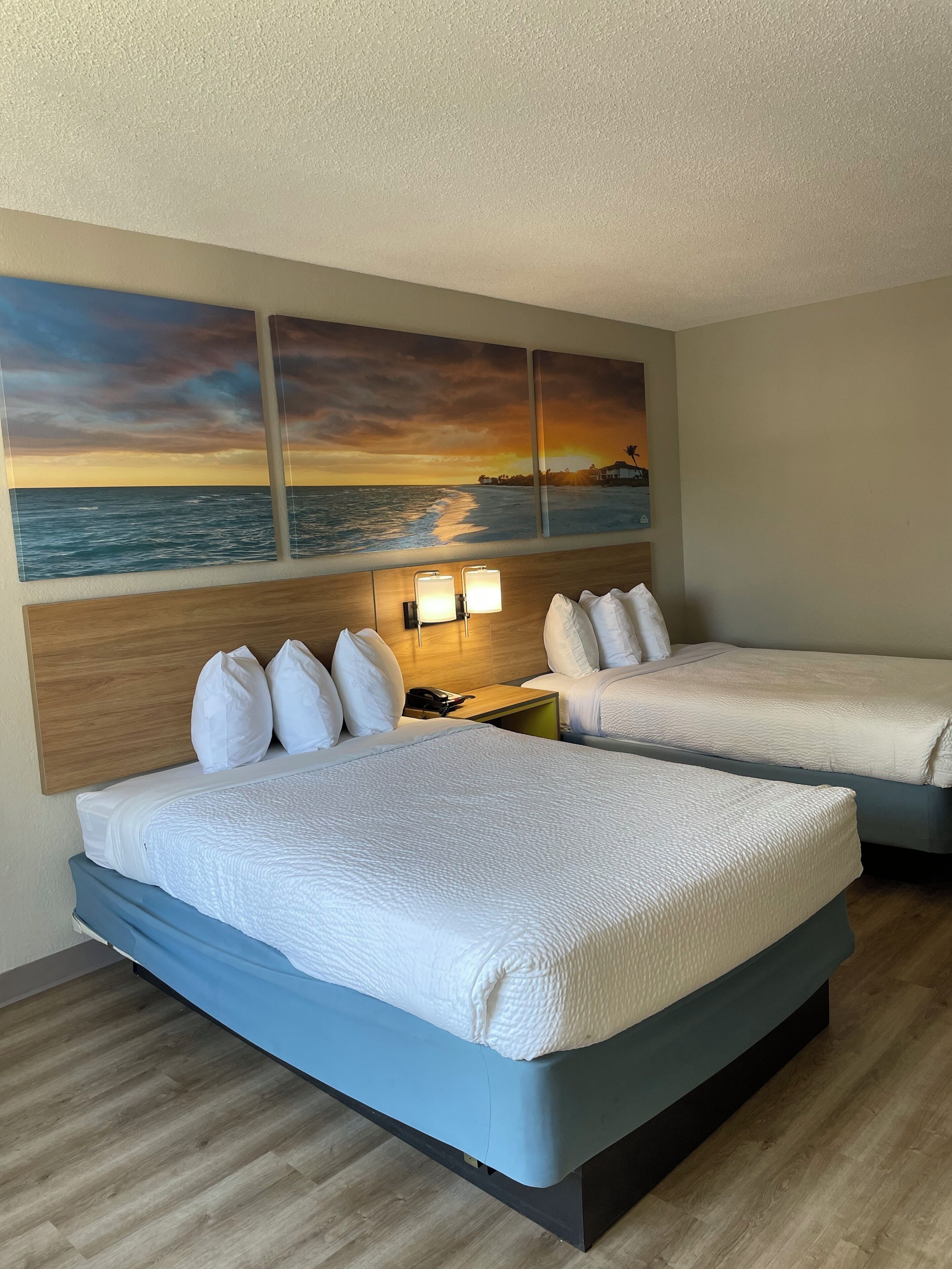 Camera d'albergo con due letti, una testiera in legno, un quadro appeso al muro raffigurante un tramonto sull'oceano e un comodino con lampade.