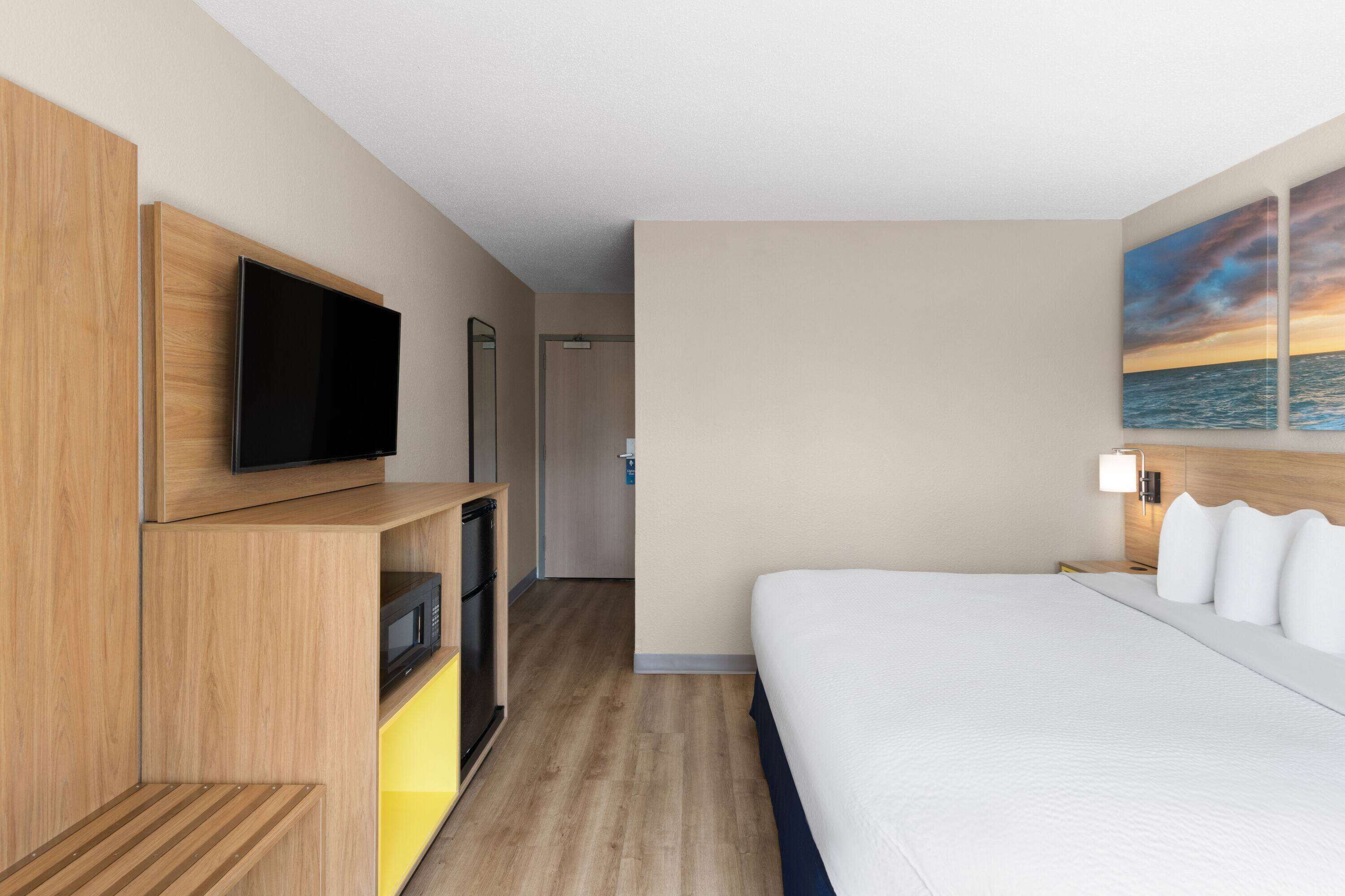 Hotelkamer met een bed, een aan een houten kast bevestigde televisie, een magnetron en een schilderij van een zonsondergang boven de oceaan.