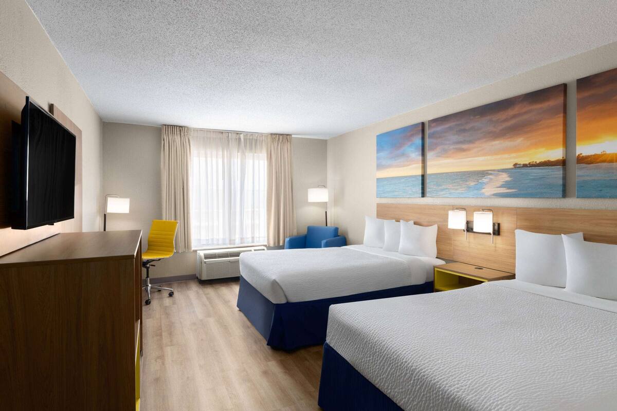 A hotel room with two beds, a desk, a chair, a TV, and a painting of a sunset over the ocean.