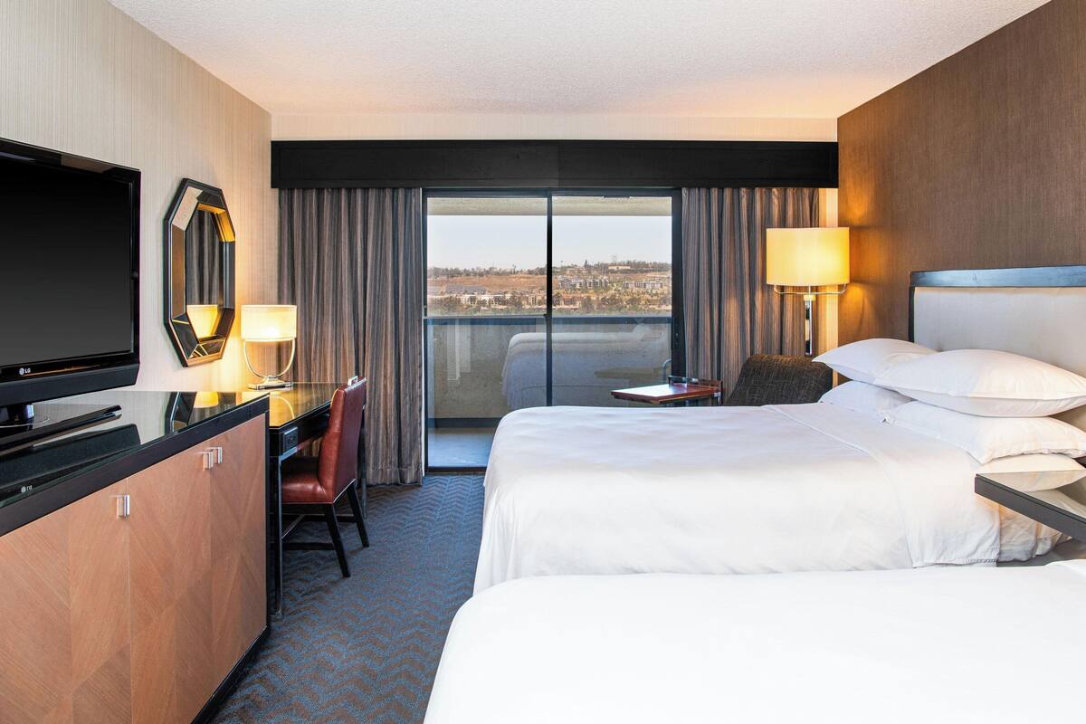 Une chambre d’hôtel avec un lit, un bureau, une télévision, une chaise, une lampe et une vue sur la ville par la fenêtre.