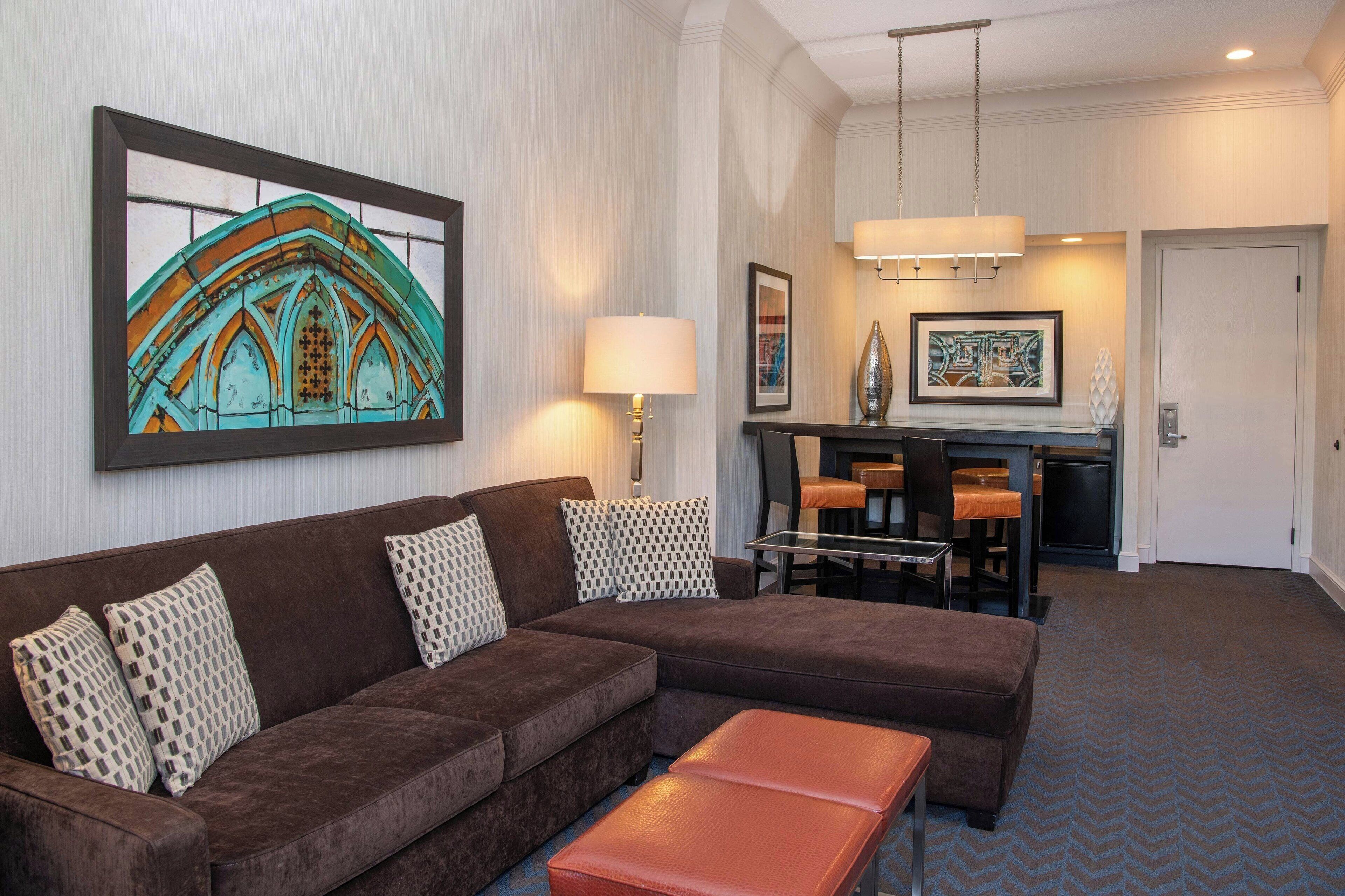 Un moderno vestíbulo de hotel con un sofá seccional marrón, una mesa de centro de vidrio, una zona de comedor con bar y obras de arte en las paredes.