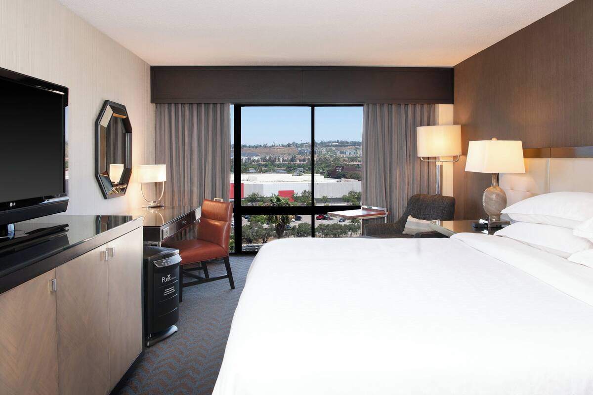 Une chambre d’hôtel dotée d’un grand lit, d’une télévision, d’un bureau avec une chaise et offrant une vue sur la ville grâce à de grandes fenêtres.