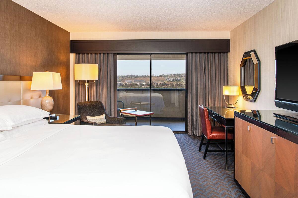 Une chambre d’hôtel avec un grand lit, un bureau avec une chaise, une télévision et une vue sur la ville par la fenêtre.