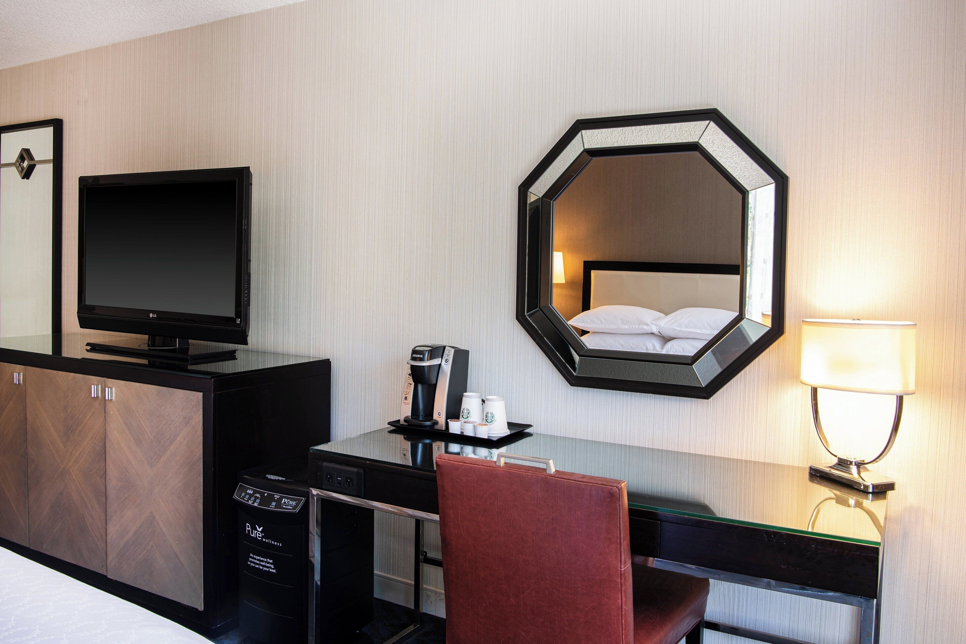 Ein Hotelzimmer mit einem Bett, einem Schreibtisch mit Computer, einem Fernseher, einer Kaffeemaschine und einem Spiegel.