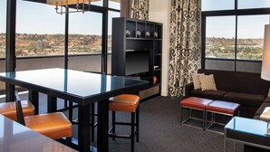 Een moderne kamer met een glazen eettafel, twee barkrukken, een bank en een tv-meubel met decoratieve objecten.