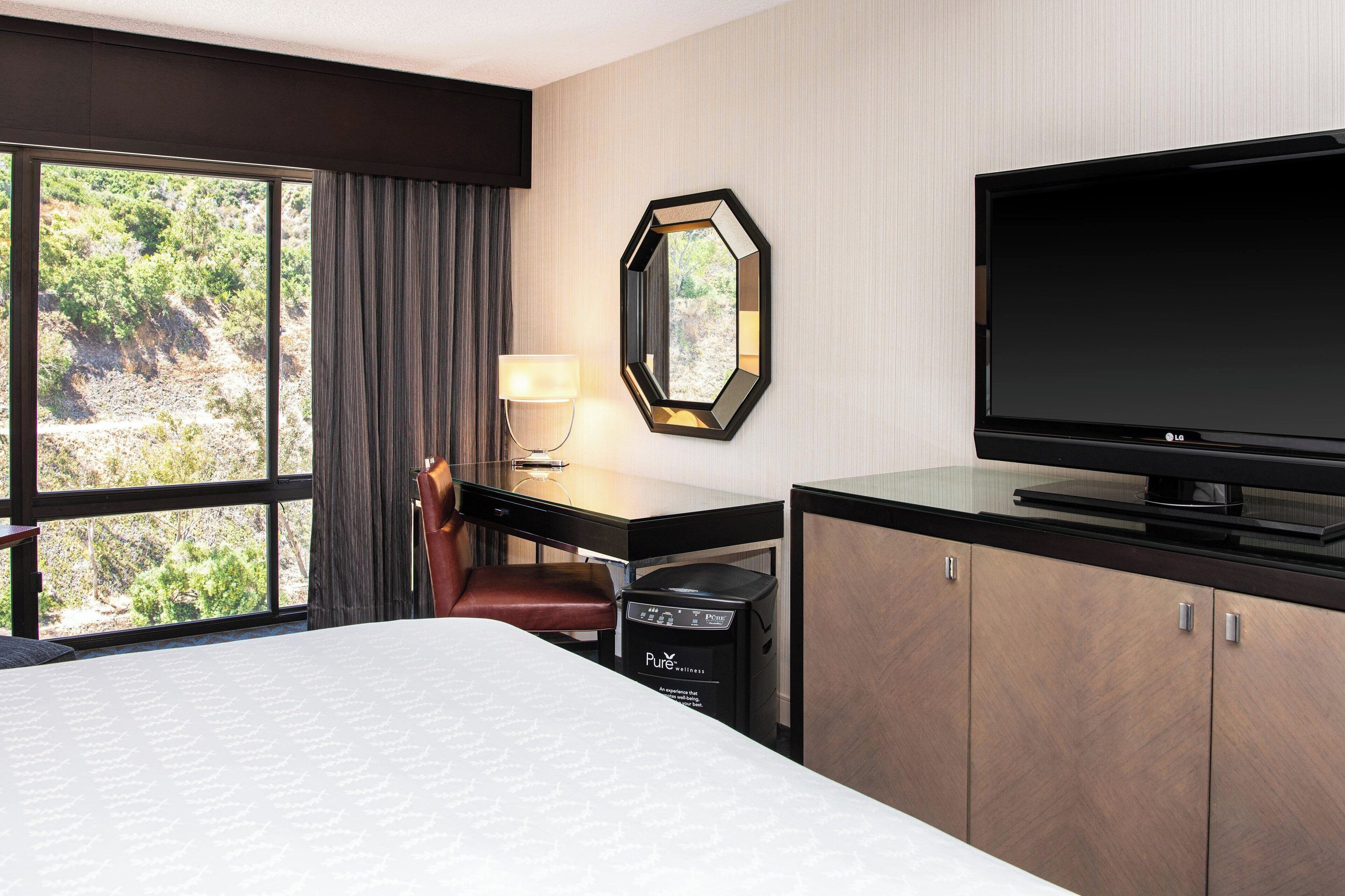 A hotel room with a bed, a desk with a chair, a TV, a mirror, and a window with curtains.