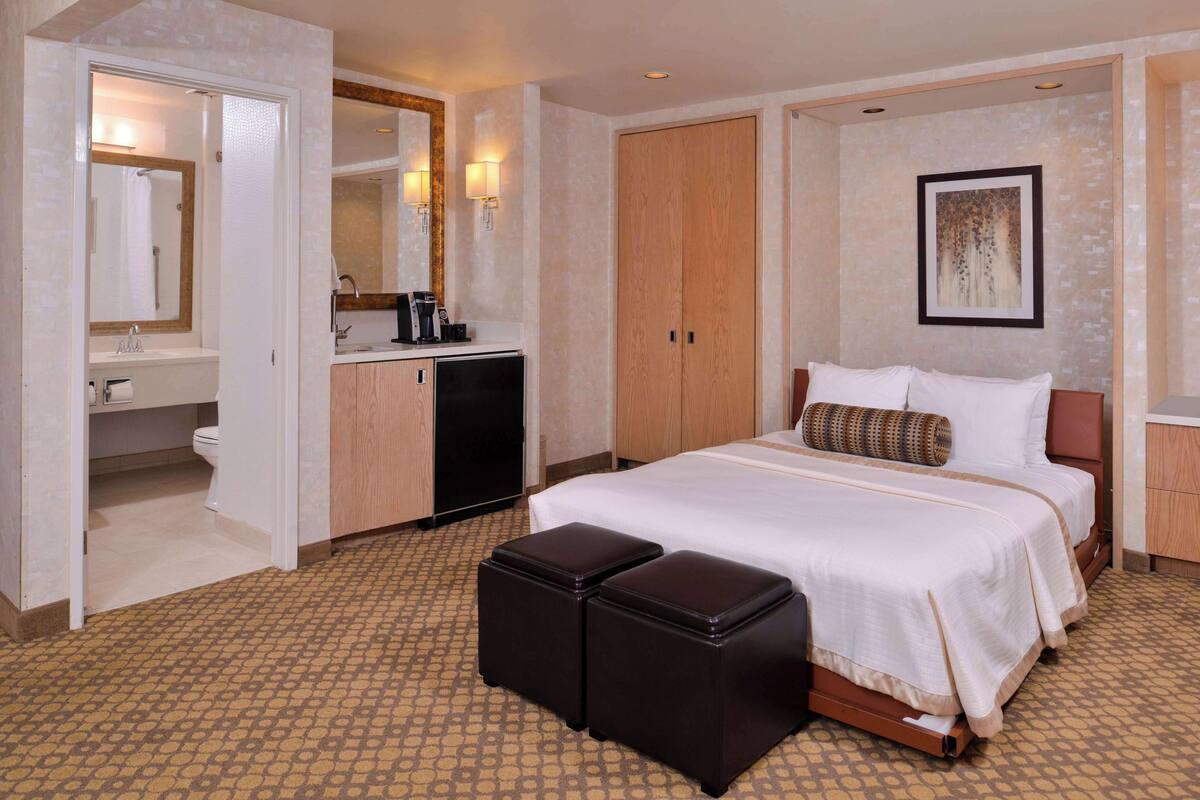 A hotel room with a large bed, a bedside table, a wardrobe, a bathroom with a sink and toilet, and a small kitchenette.
