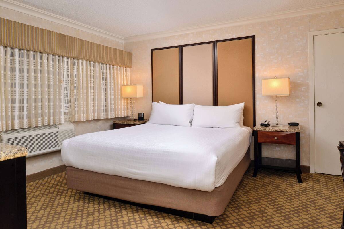 A hotel room with a large bed, two bedside tables, a window with curtains, and a door.