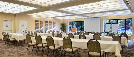 Un salón de conferencias con mesas largas y sillas, una pizarra y un ventanal con vistas a árboles.