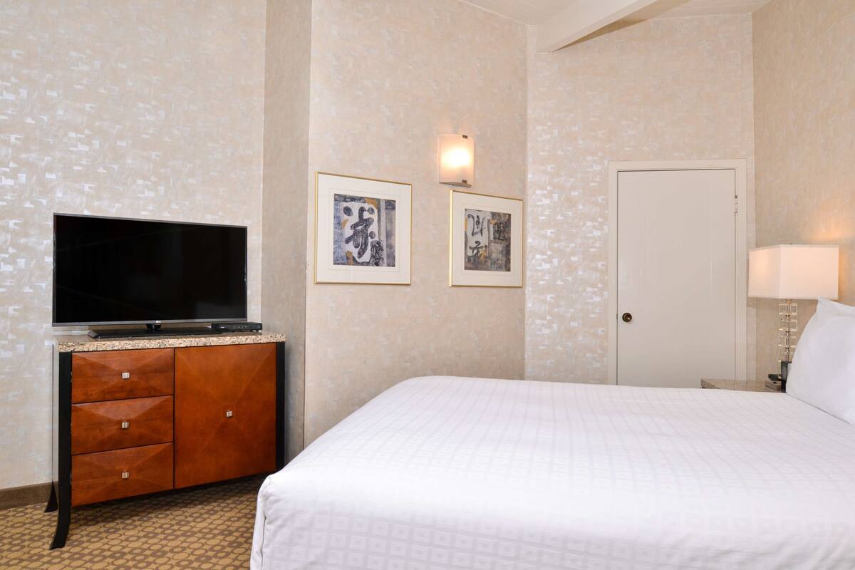 A hotel room with a bed, a television on a wooden cabinet, two framed calligraphy artworks, a door, and a wall-mounted light.