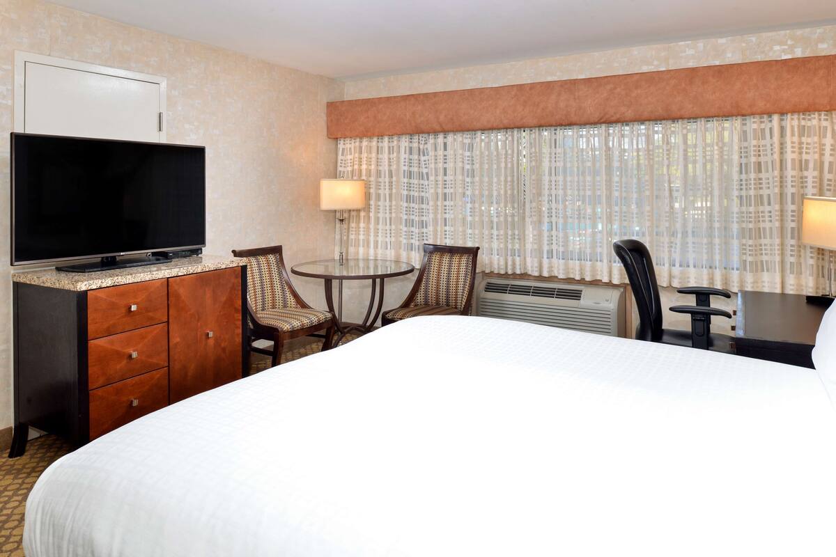 A hotel room with a bed, a TV, a desk, two chairs, and a window with curtains.