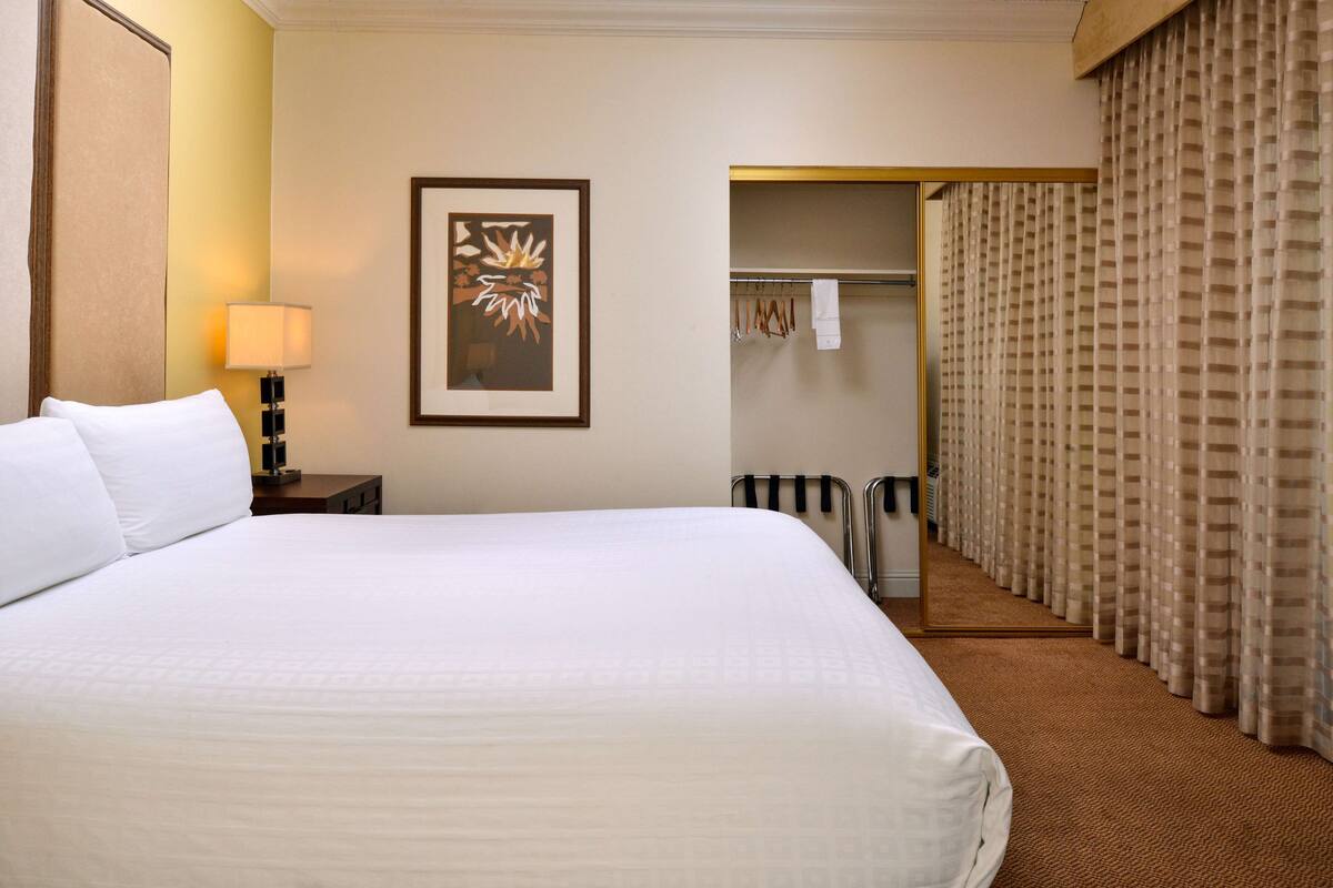 A hotel room with a large bed, a nightstand with a lamp, a framed picture on the wall, a closet with hanging clothes, and a bathroom with a towel rack.