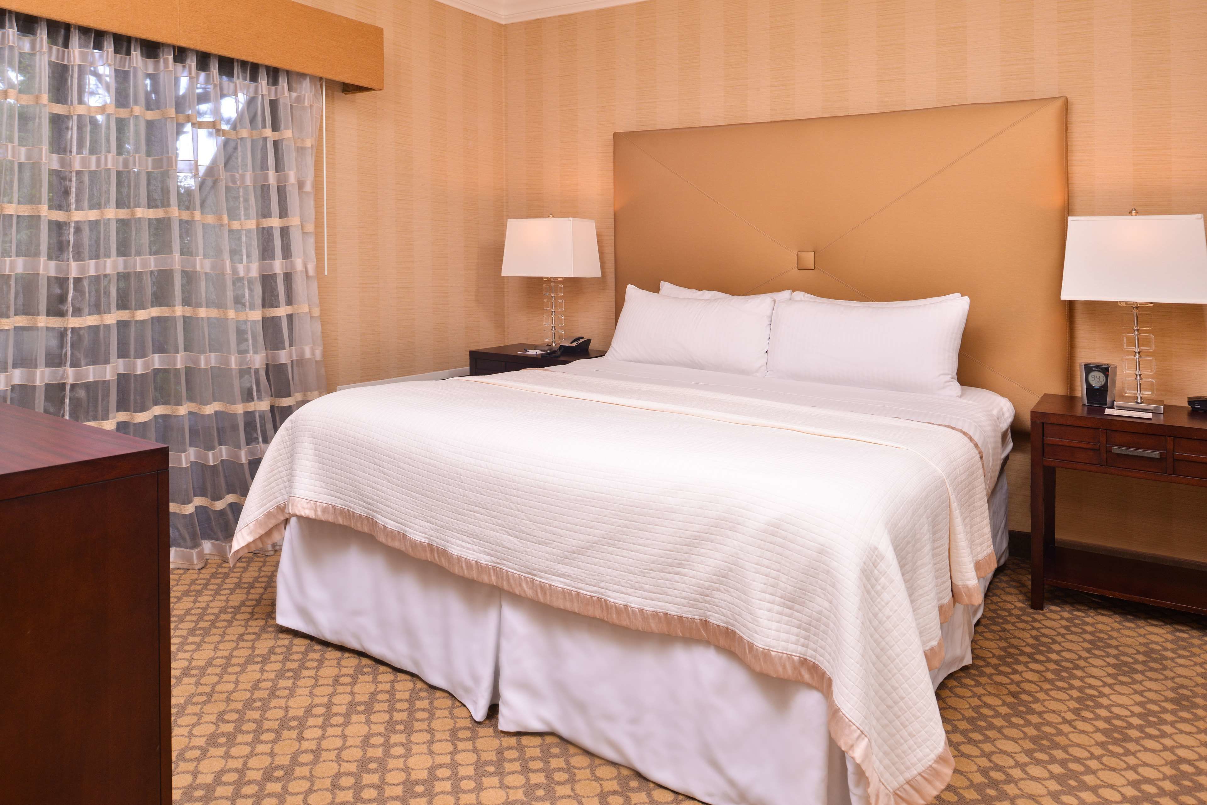 A hotel room with a large bed, two bedside tables, a nightstand, a wooden desk, and a patterned carpet.