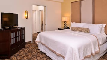 Quarto de hotel com uma cama grande, uma televisão sobre um móvel de madeira, uma mesa de cabeceira com abajur e um tapete estampado.