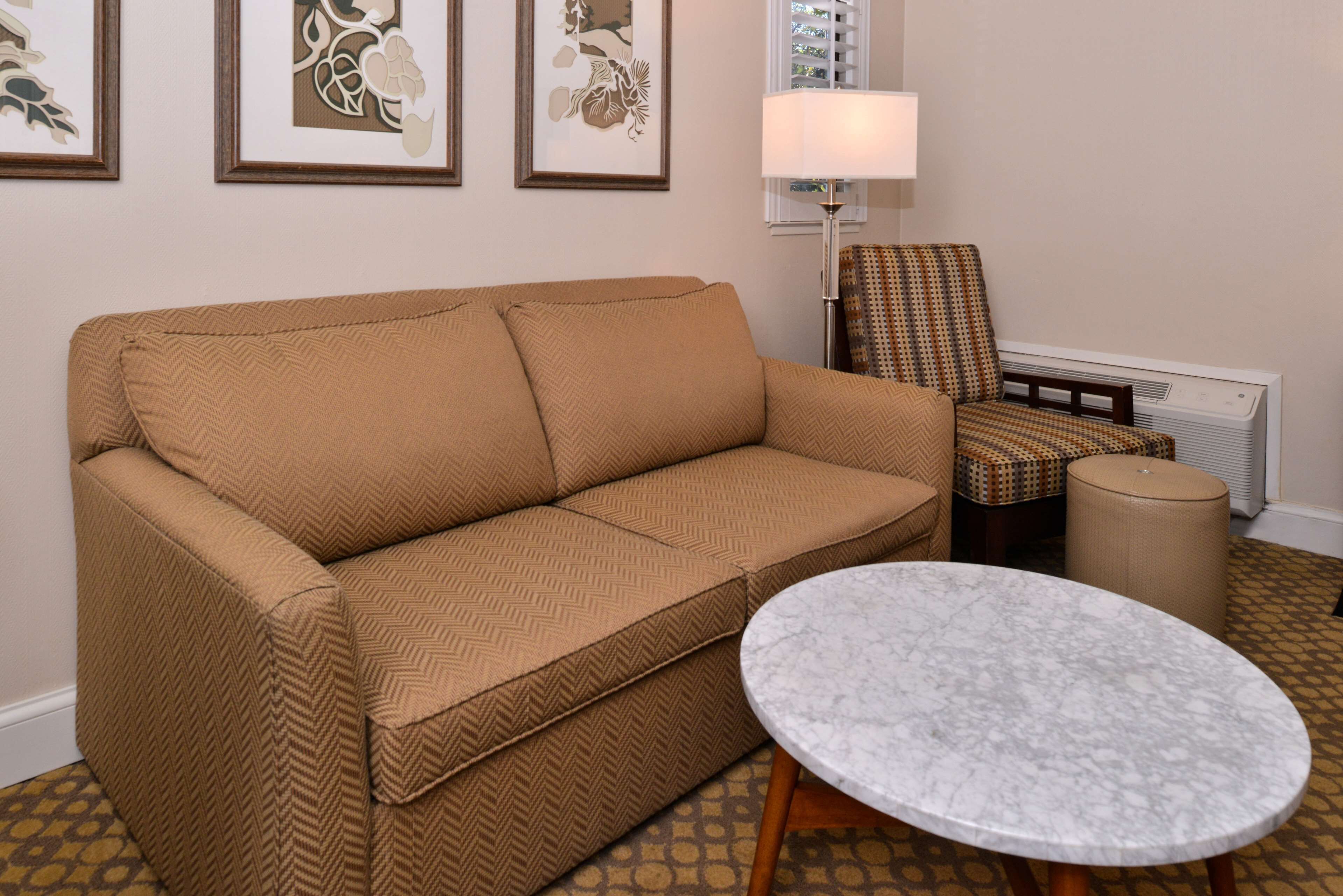 A beige sofa, a patterned armchair, a round marble table, and a floor lamp in a room with patterned carpet.