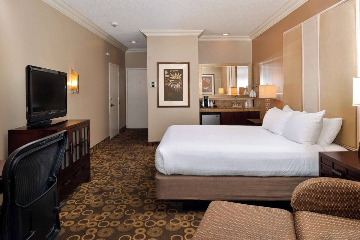 A hotel room with a bed, a TV, a desk, a chair, a bathroom with a sink and mirror, and a patterned carpet.