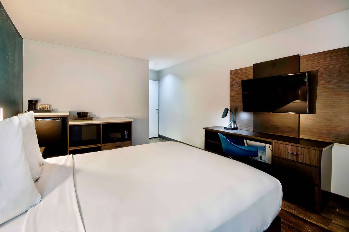 Ein modernes Hotelzimmer mit einem großen Bett, einem Schreibtisch mit Flachbildfernseher, einem Sessel und einer kleinen Küchenzeile.