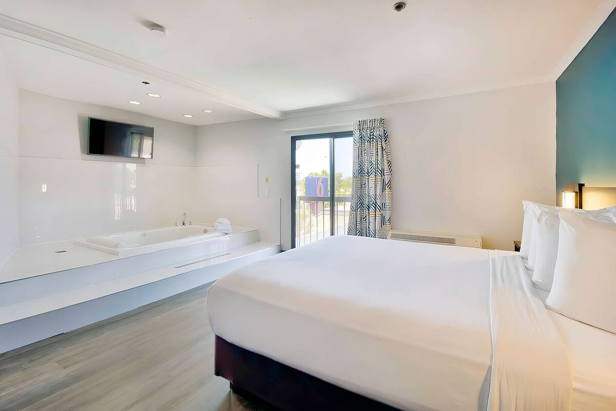Ein modernes Hotelzimmer mit einem großen Bett, einer integrierten Badewanne, einem Flachbildfernseher und einem großen Fenster mit Außenblick.
