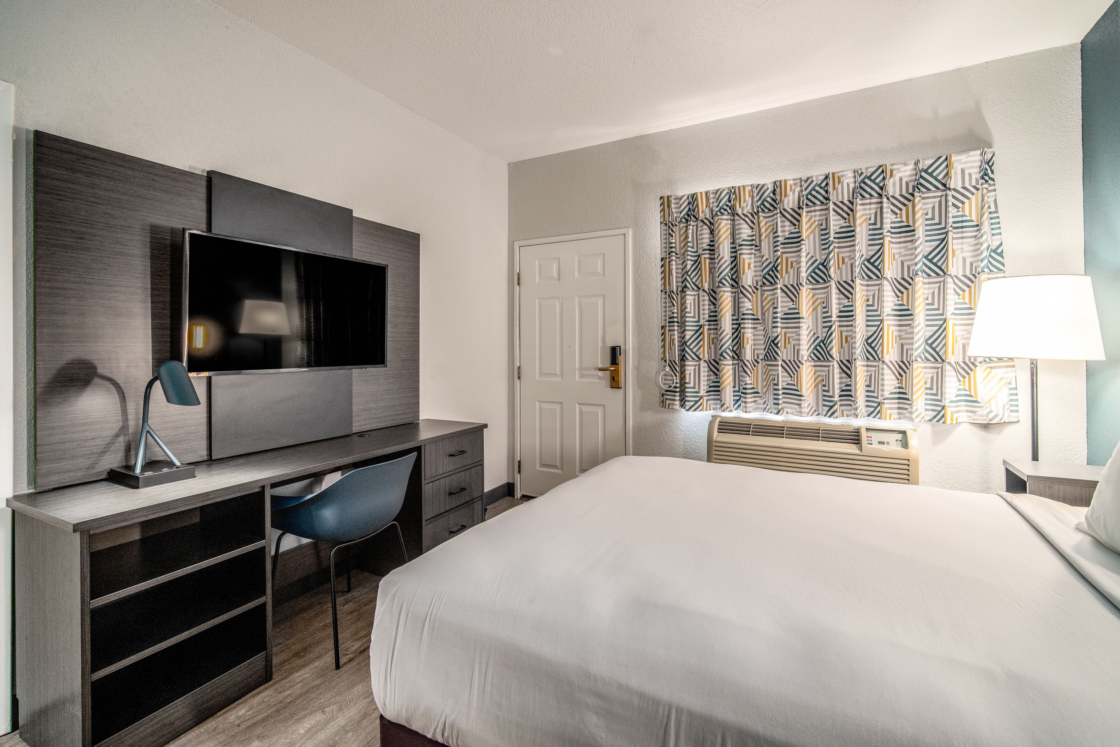A hotel room with a bed, a desk with a chair, a television, and a patterned curtain.