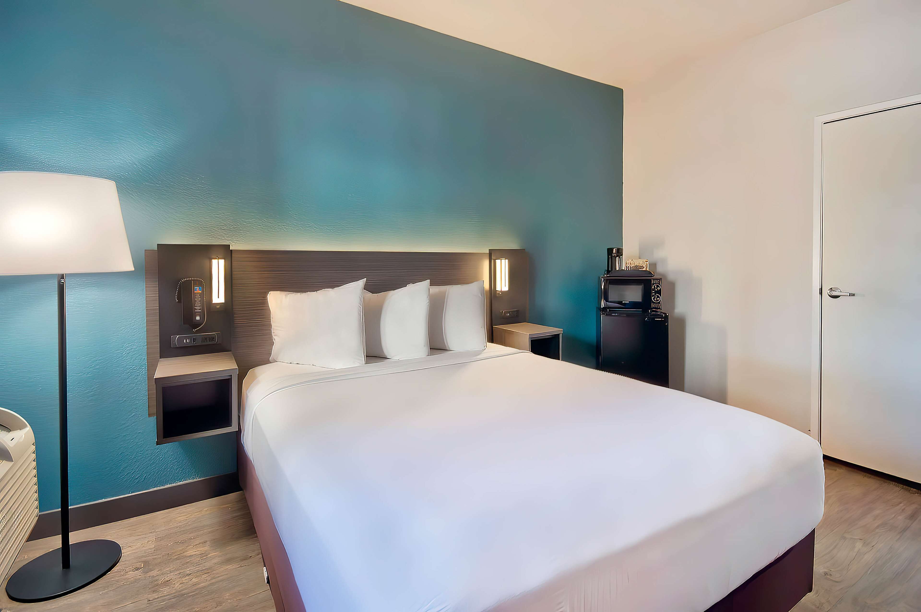 A hotel room with a large bed, bedside tables, a lamp, a phone, and a small shelf with items.