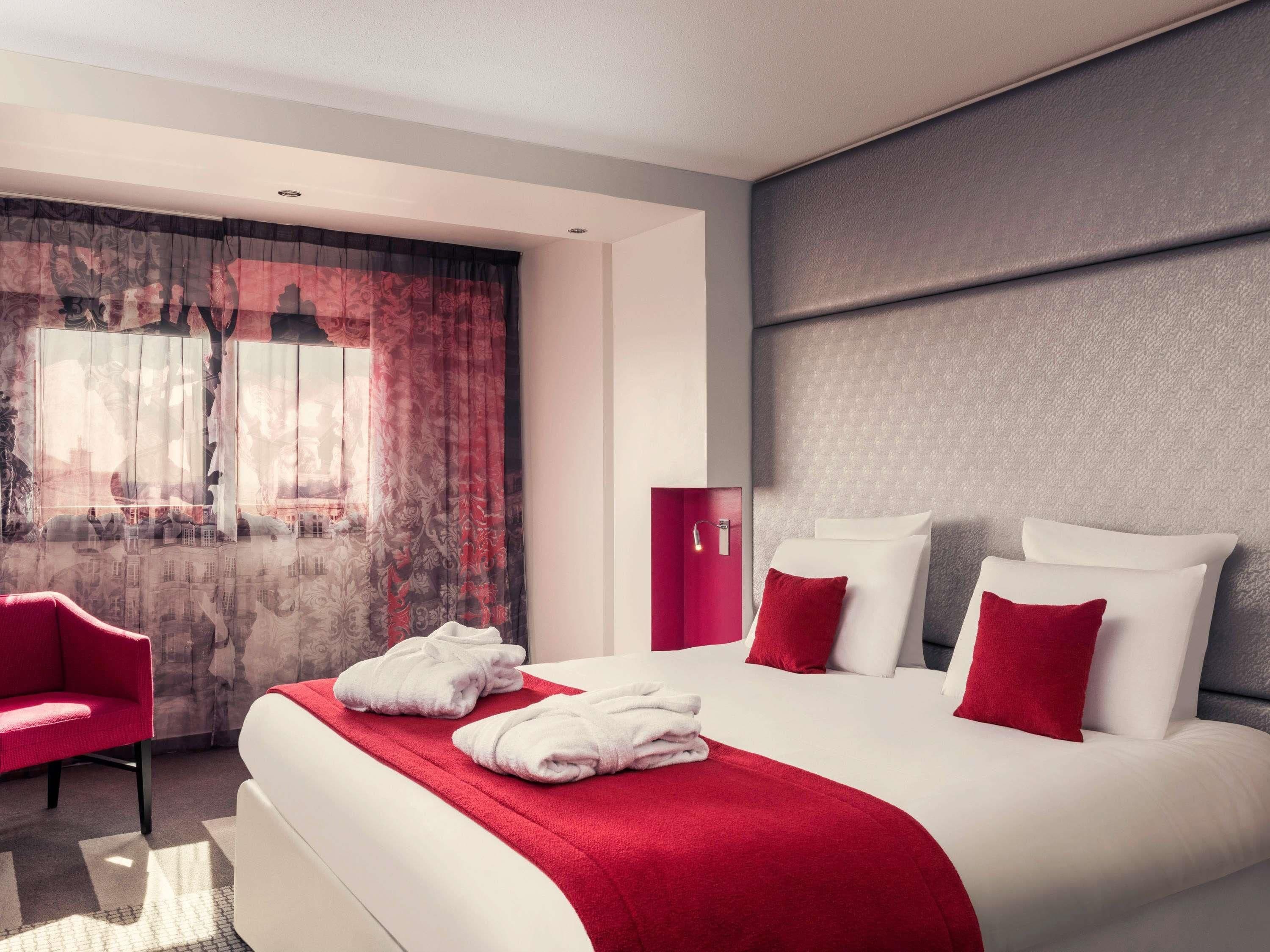 A modern hotel room with a large bed, red and white bedding, a red chair, and a view of a cityscape through the window.