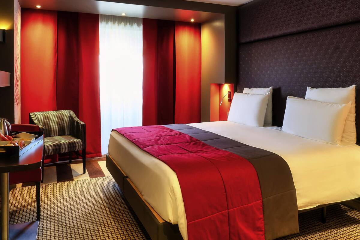 A hotel room with a large bed, red curtains, a desk with a chair, and a patterned floor.