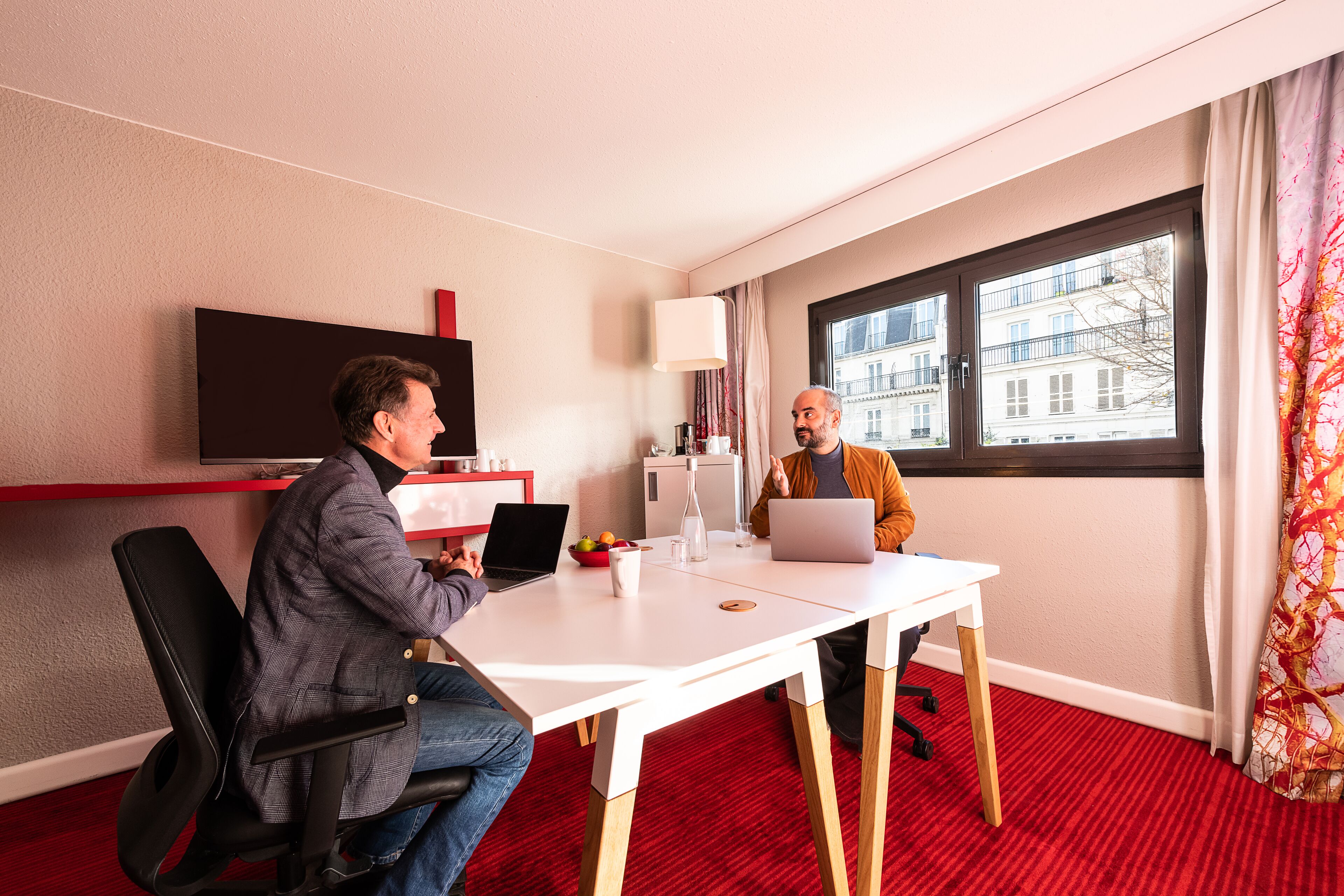 A hotel room with a red carpet, a white desk, two laptops, a flat-screen TV, and a window with a view of buildings.