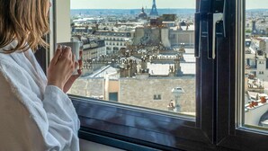 Een persoon in een witte badjas met een glas in de hand, kijkt uit het raam op een stadsgezicht met de Eiffeltoren.