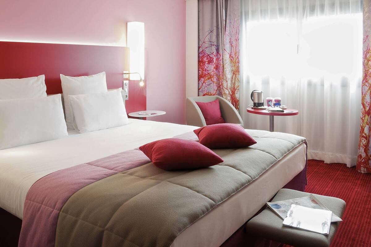 A hotel room with a large bed, a bedside table, a chair, a small table with a kettle, and a window with curtains.