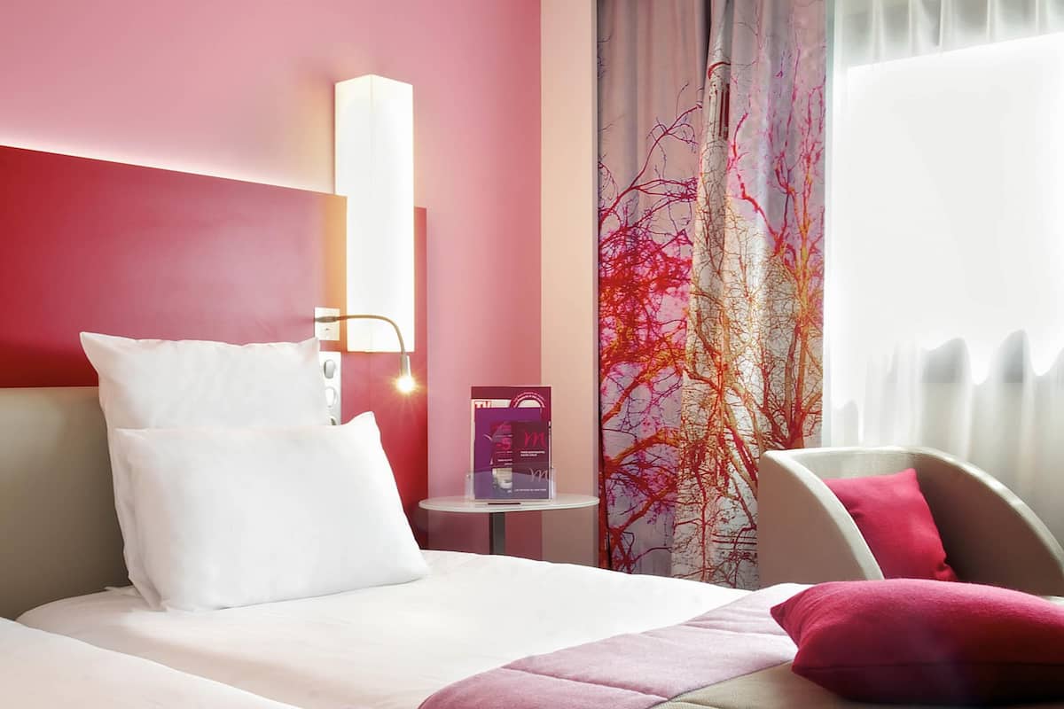 Ein Hotelzimmer mit einem Bett, Kissen, einem Nachttisch, einer Lampe, einem Fenster mit Vorhängen und einer Wand mit einem Baumdesign.