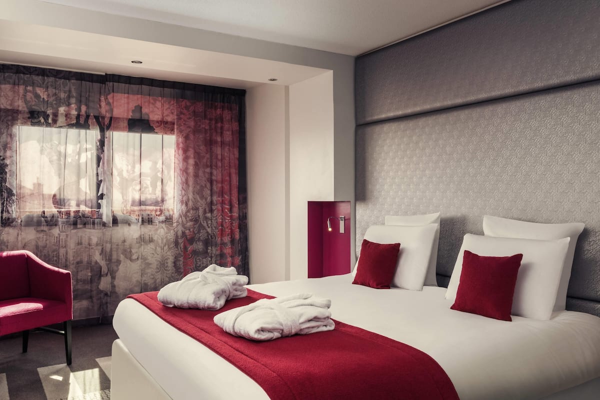 Una habitación de hotel moderna con una cama grande, almohadas rojas y blancas, una silla roja y vistas a un paisaje urbano por la ventana.
