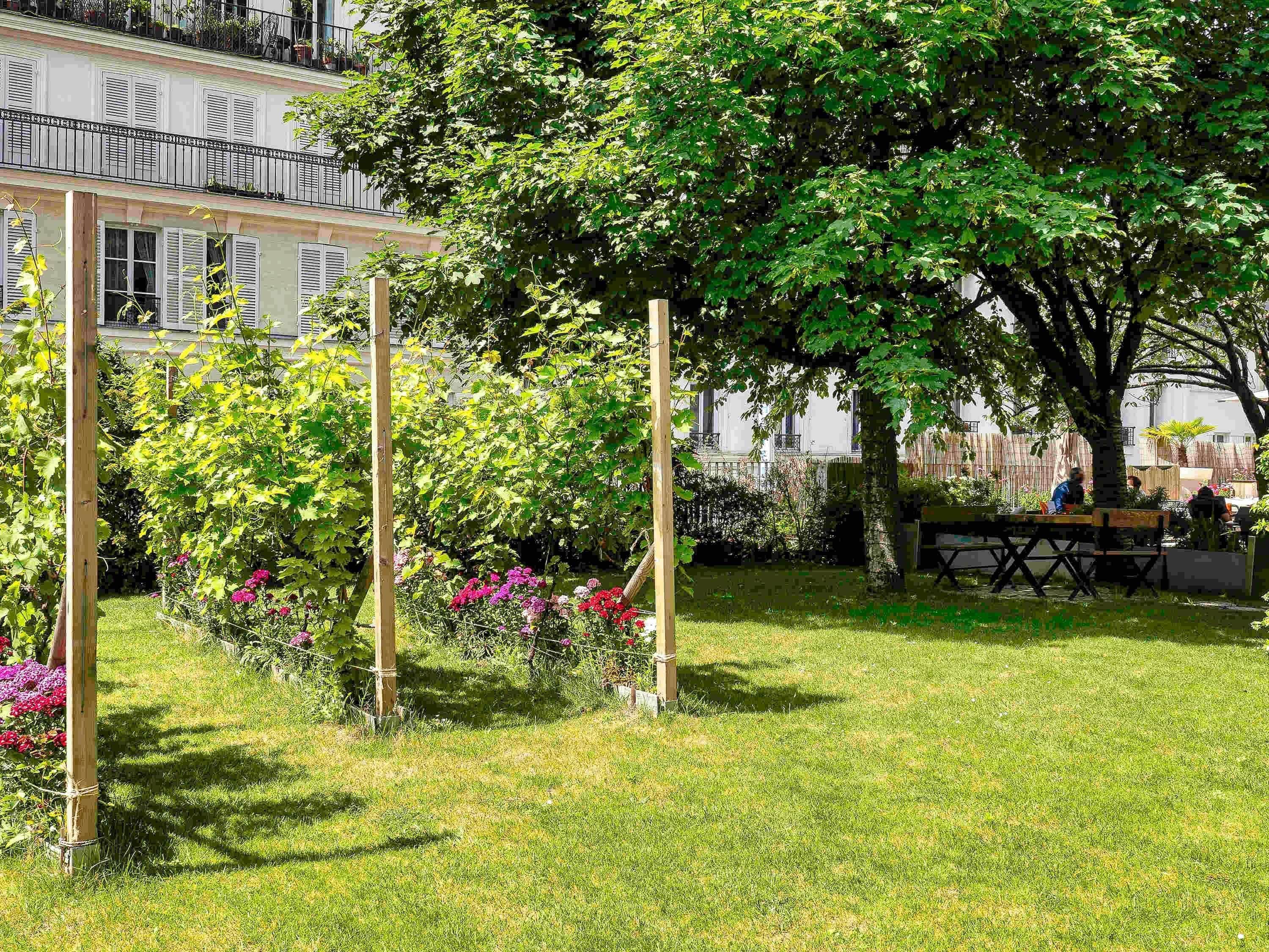 A garden with a wooden pergola, a table with chairs, and a building with balconies in the background.