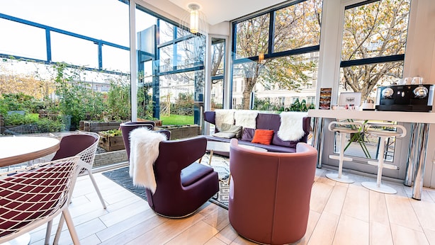 A modern outdoor seating area with a glass wall, a bar with stools, and a view of the garden. - Mercure Paris Montmartre Sacré Coeur (Paris)