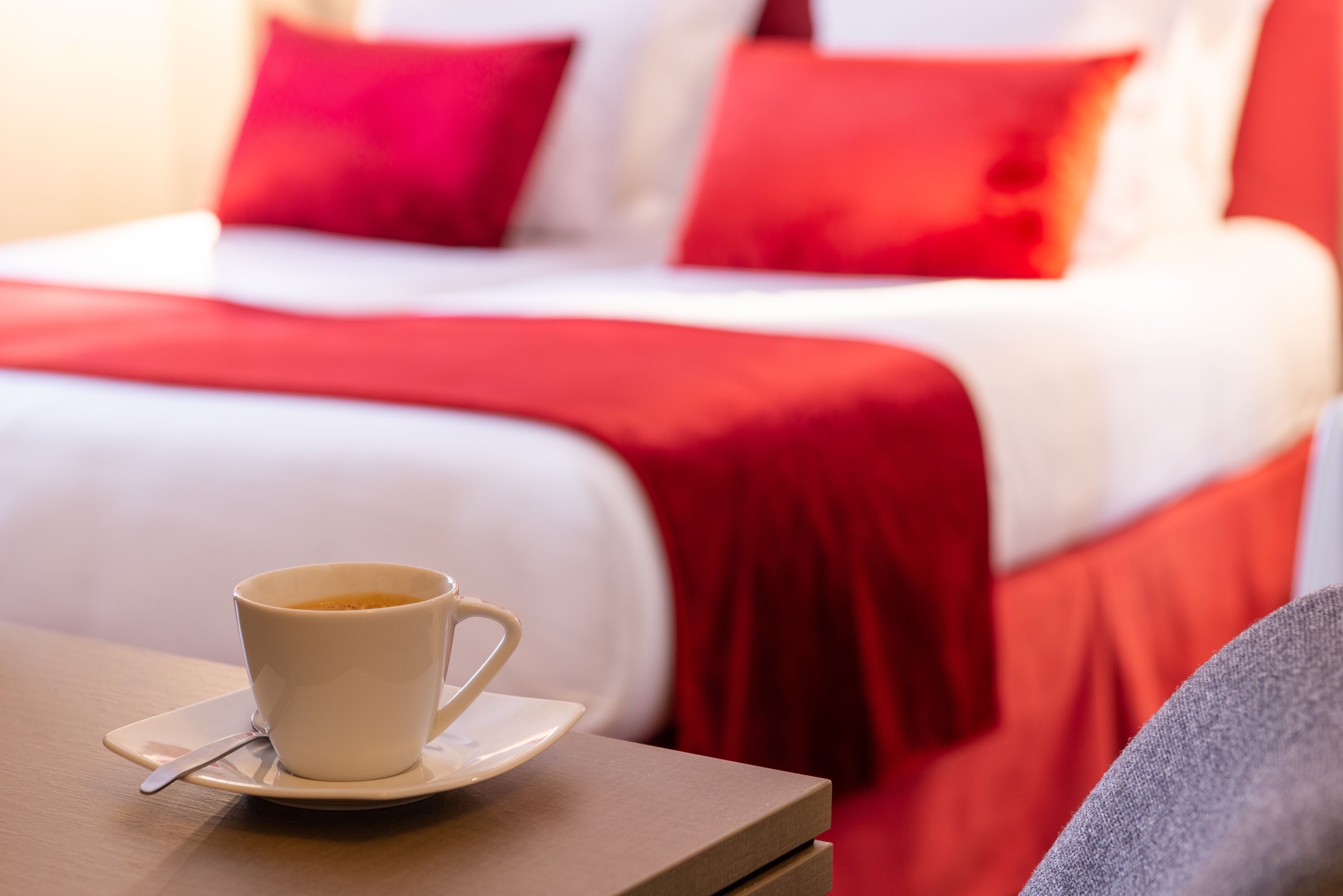 A hotel room with a bed, a cup of coffee on a table, and red bedding.