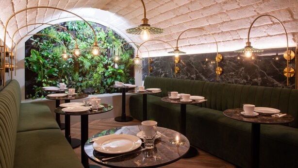 Un restaurant avec des tables rondes, des sièges recouverts d’un tissu vert et un mur en briques agrémenté de plantes.
