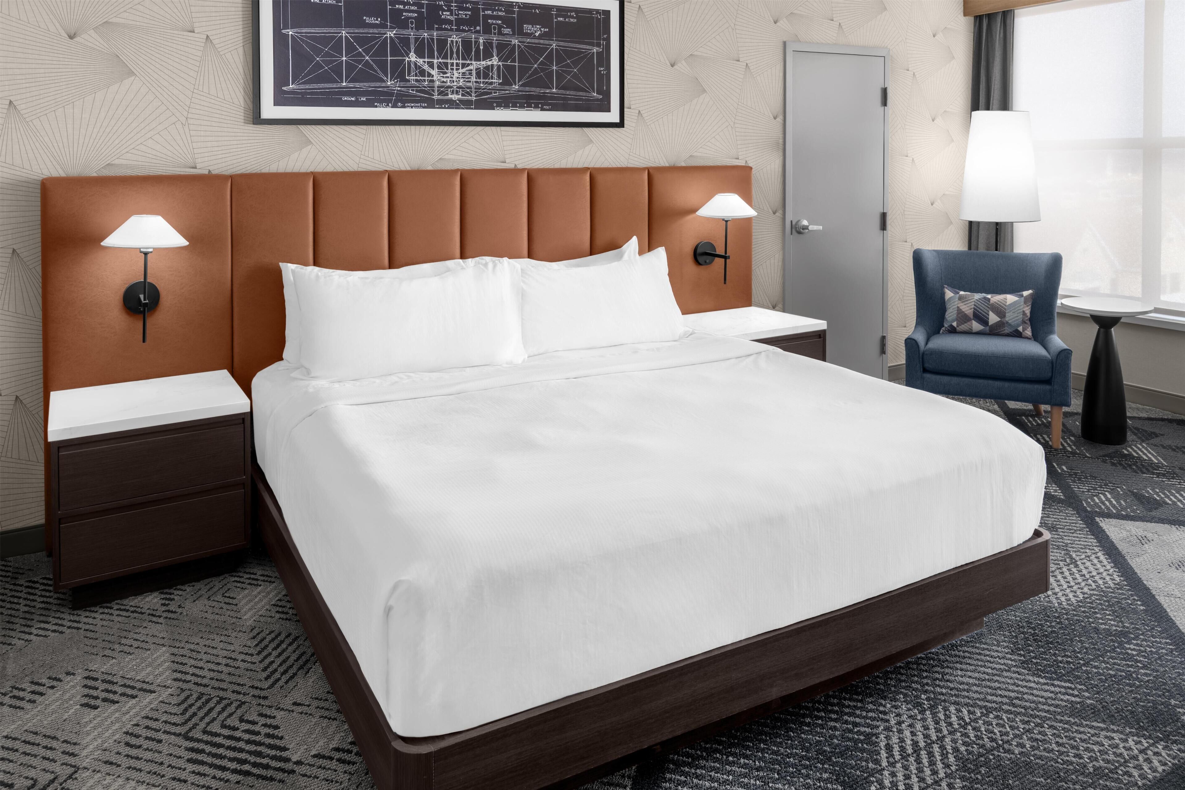 Een hotelkamer met een groot bed, twee nachtkastjes, een stoel, een lamp en een ingelijst architectuurtekening aan de muur.