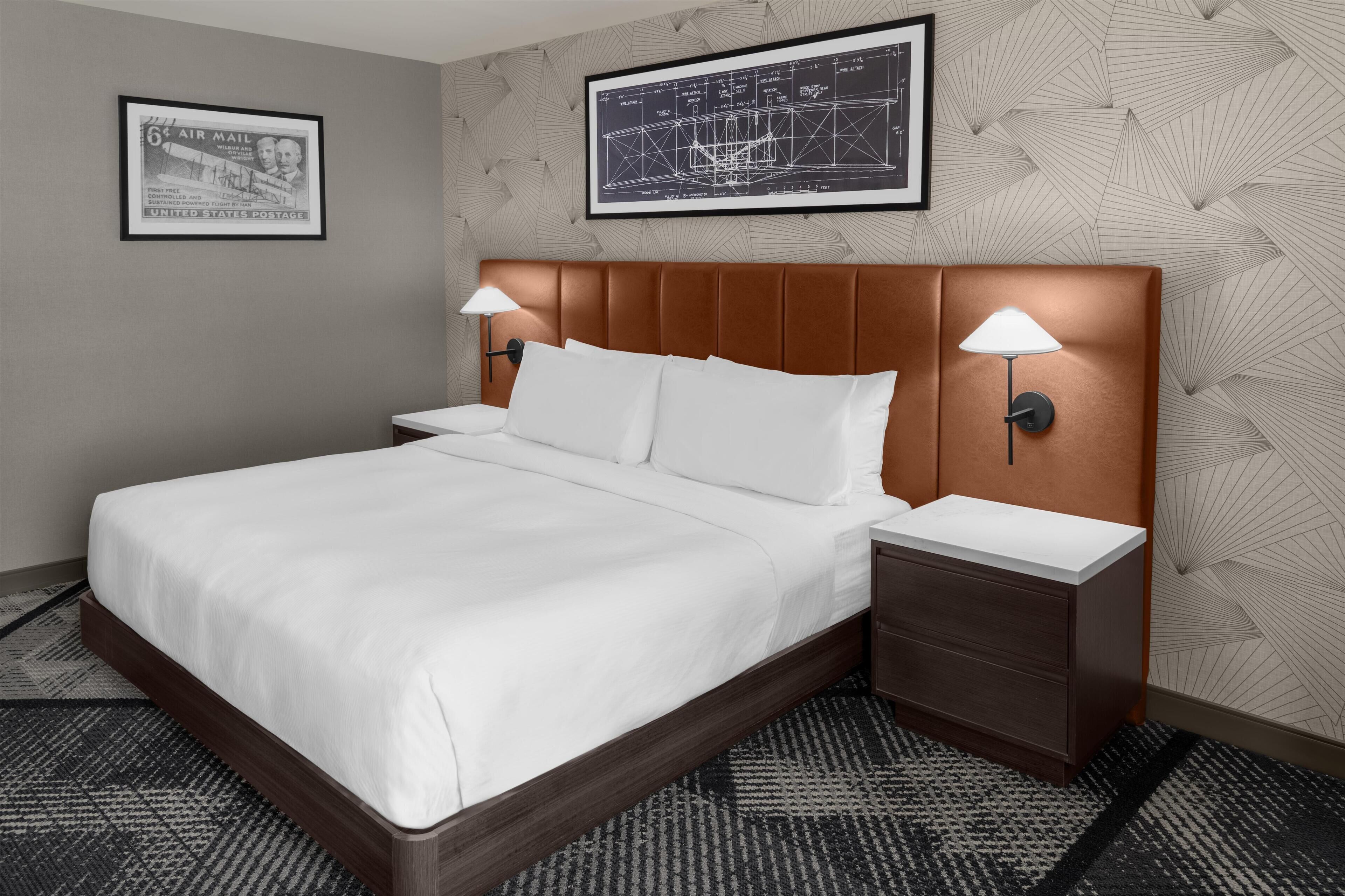 A hotel room with a large bed, two bedside tables with lamps, a headboard, a framed map on the wall, and a patterned carpet.