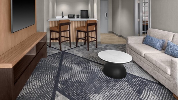 A modern living room with a flat-screen TV, a sofa, a coffee table, and a kitchen area with a bar stool. - DoubleTree by Hilton Dayton Fairborn (Fairborn)