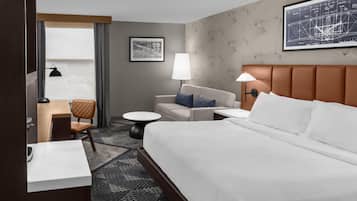 A hotel room with a large bed, a sofa, a desk, and a chair. There is a window with curtains, a wall-mounted map, and a patterned wall.