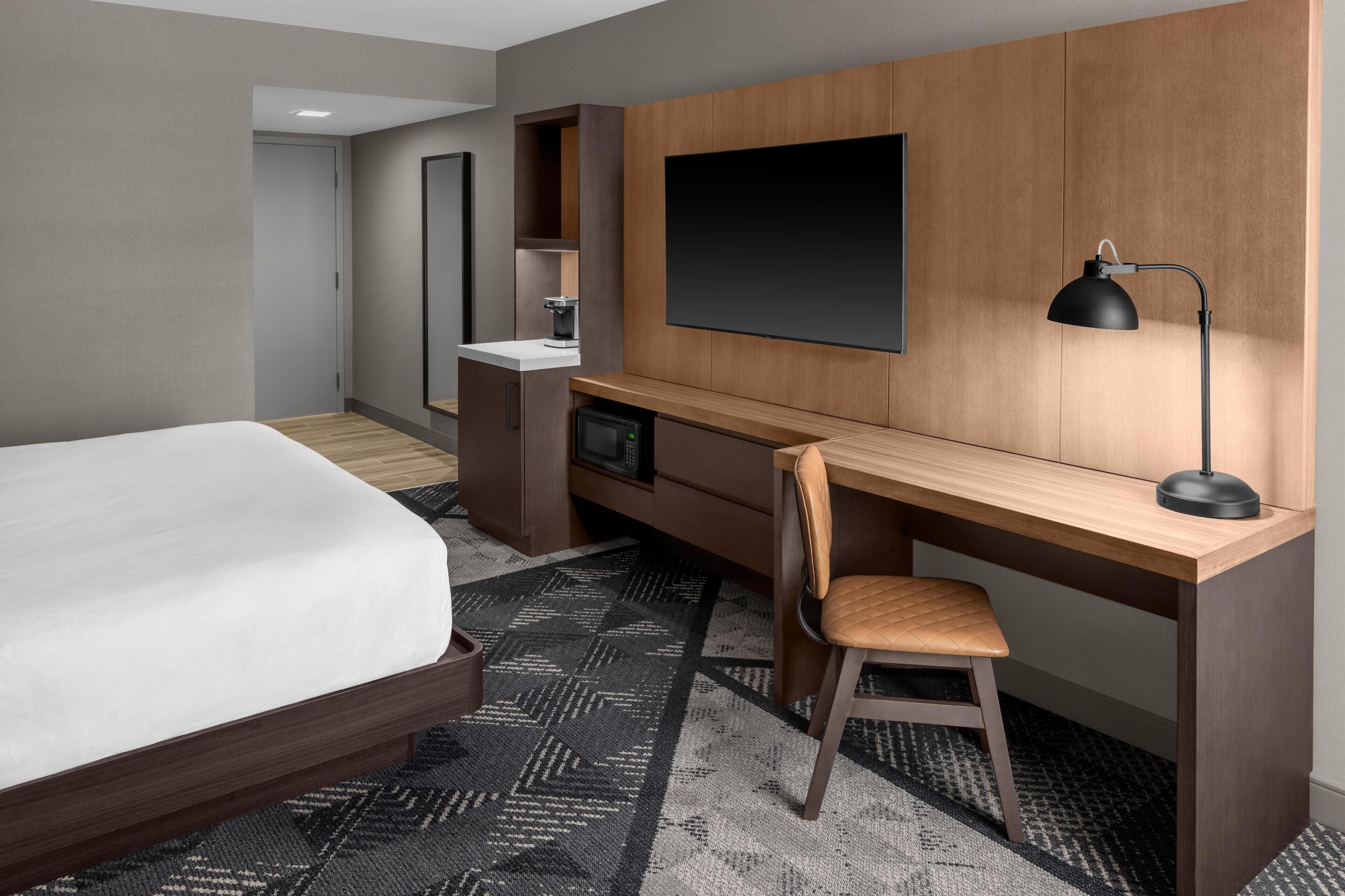 Une chambre d’hôtel comprenant un lit, un bureau avec une chaise, une télévision, une machine à café et un tapis à motifs.