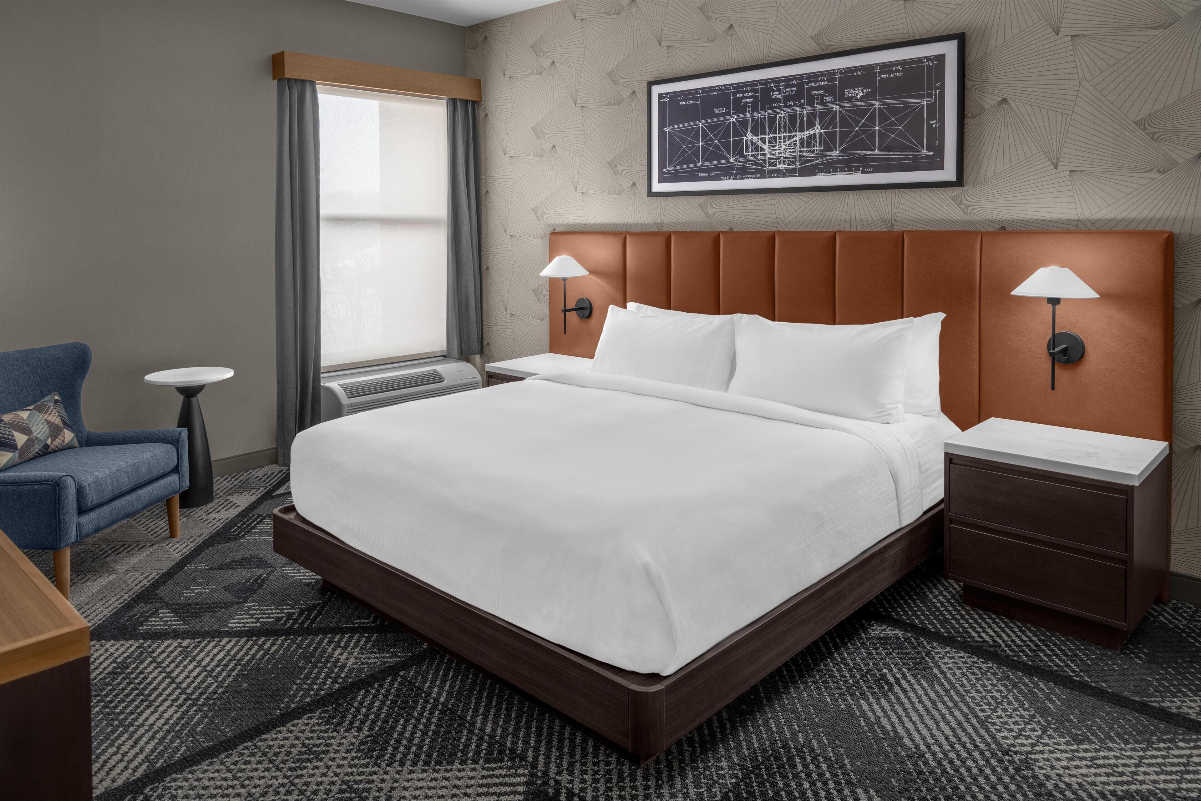 A hotel room with a large bed, two bedside tables with lamps, a chair, a window with curtains, and a framed architectural drawing on the wall.