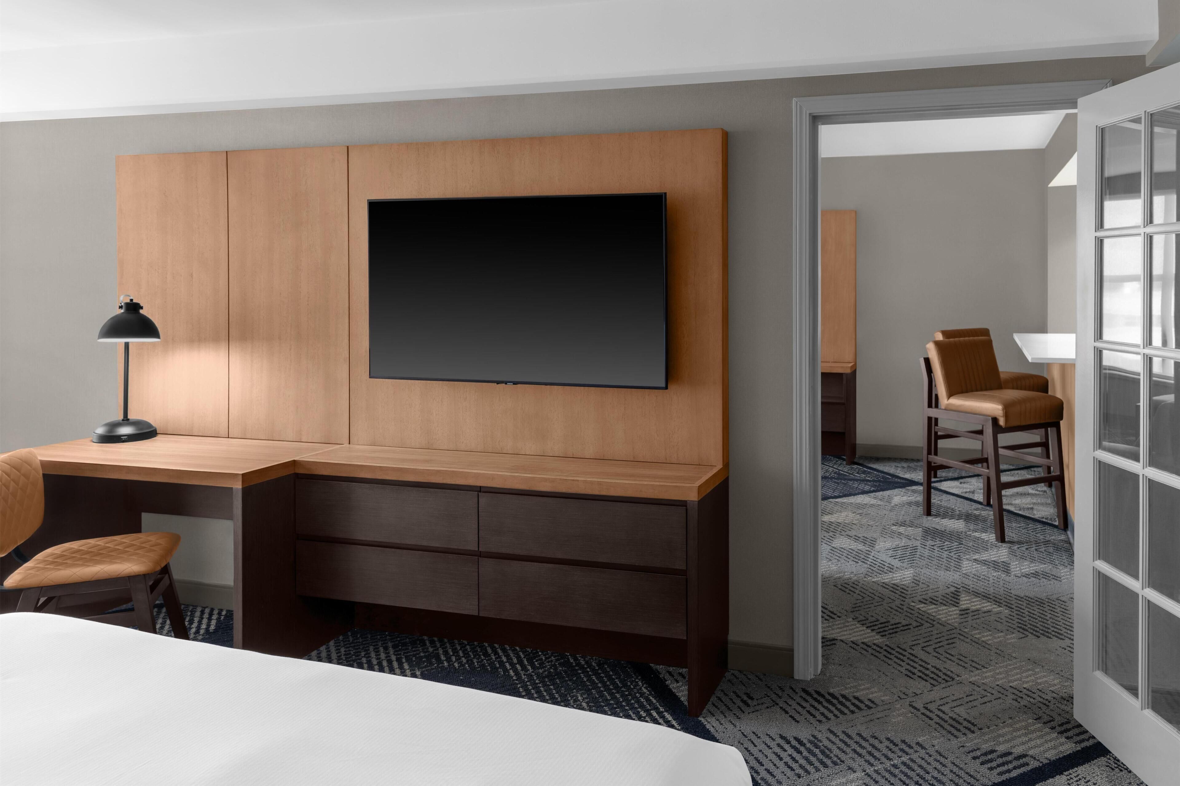 Habitación de hotel con un escritorio de madera, un televisor de pantalla plana montado en la pared, una silla, una lámpara y una alfombra estampada.